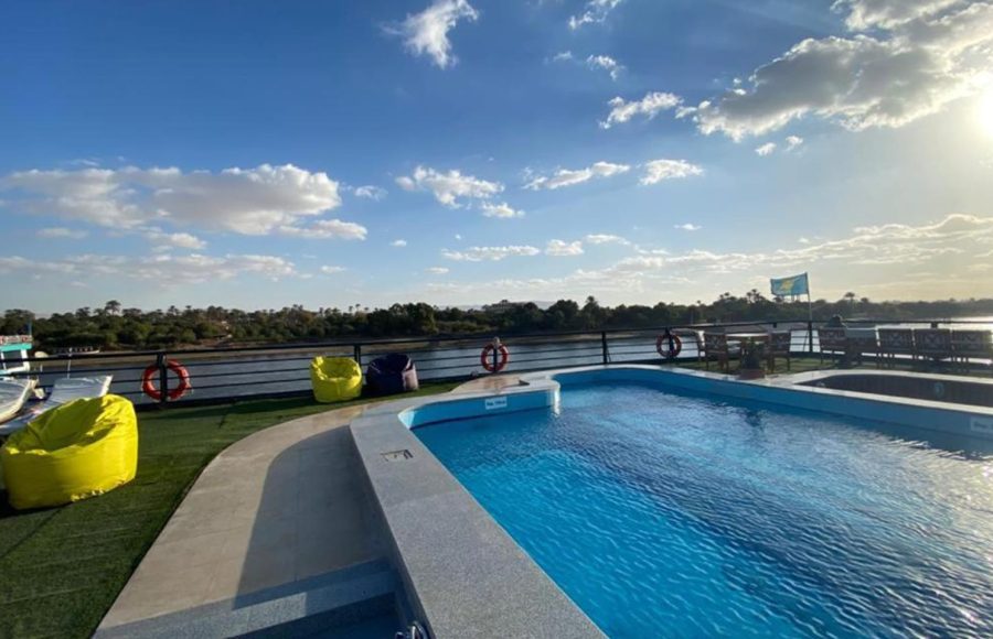 The sun deck of a Nile Cruise ship with a rectangular swimming pool, bordered by a synthetic green lawn and bright yellow and blue beanbag chairs, under a clear blue sky.