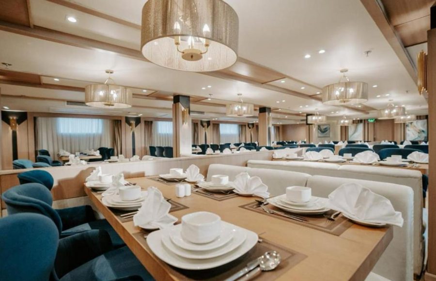 The elegant main restaurant of a Nile Cruise ship, featuring a long table set with white china in the foreground, surrounded by deep blue velvet chairs and light-colored booth seating, illuminated by contemporary drum chandeliers.