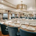 The main dining hall of a Nile Cruise ship, featuring long tables set with white china, deep blue velvet dining chairs, booth seating, and contemporary gold mesh drum chandeliers.