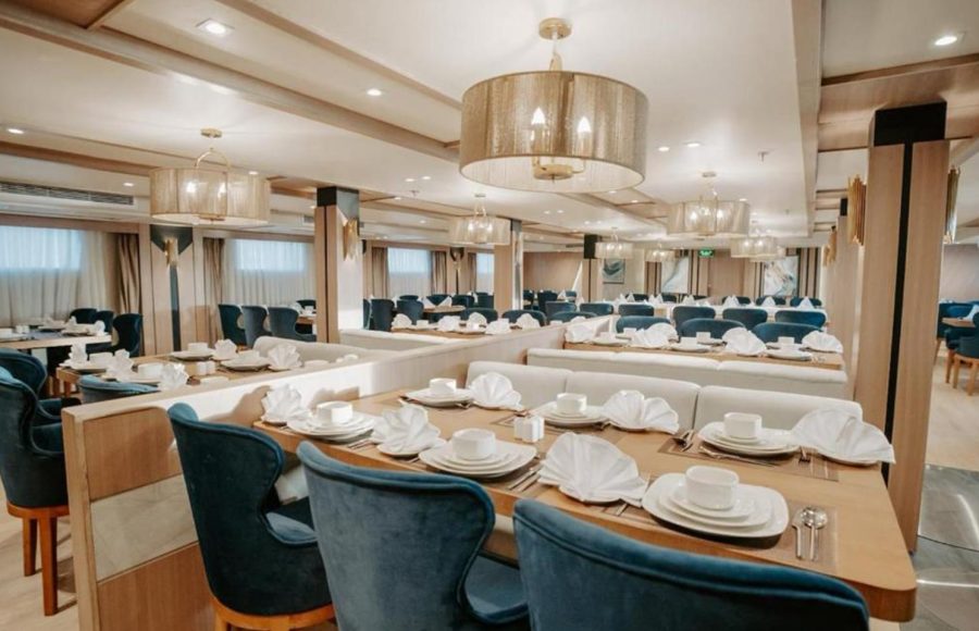 The main dining hall of a Nile Cruise ship, featuring long tables set with white china, deep blue velvet dining chairs, booth seating, and contemporary gold mesh drum chandeliers.