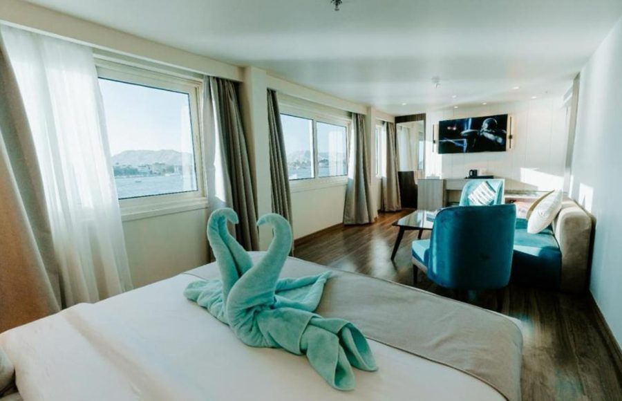 Bright, modern Nile Cruise cabin featuring a double bed with towel swans made from light blue towels, large windows offering a river view, and a small sitting area with a couch and teal accent chair.