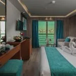 A luxurious Nile Cruise cabin featuring a large bed with a turquoise runner, towel swans, and a sliding glass door leading to a private balcony with a river view.