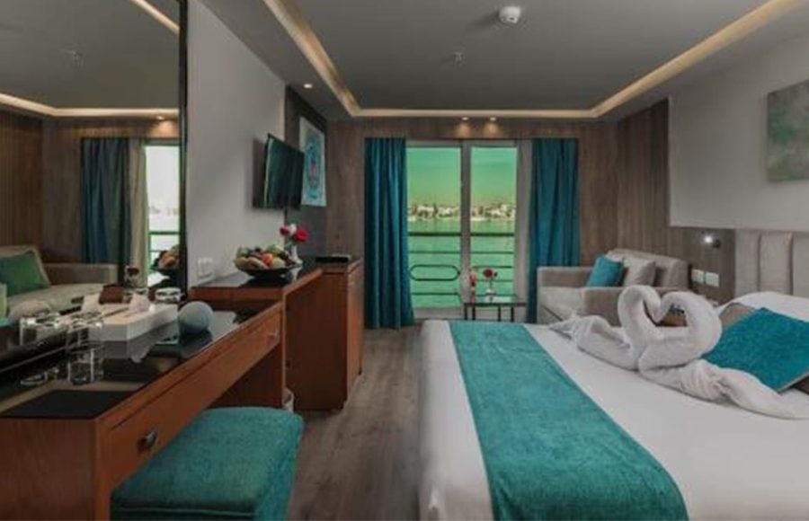 A luxurious Nile Cruise cabin featuring a large bed with a turquoise runner, towel swans, and a sliding glass door leading to a private balcony with a river view.