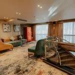 A contemporary lounge area on a Nile Cruise ship with a spiral staircase, featuring a patterned carpet, modern furniture in gold and deep green velvet, and large windows with heavy drapes.
