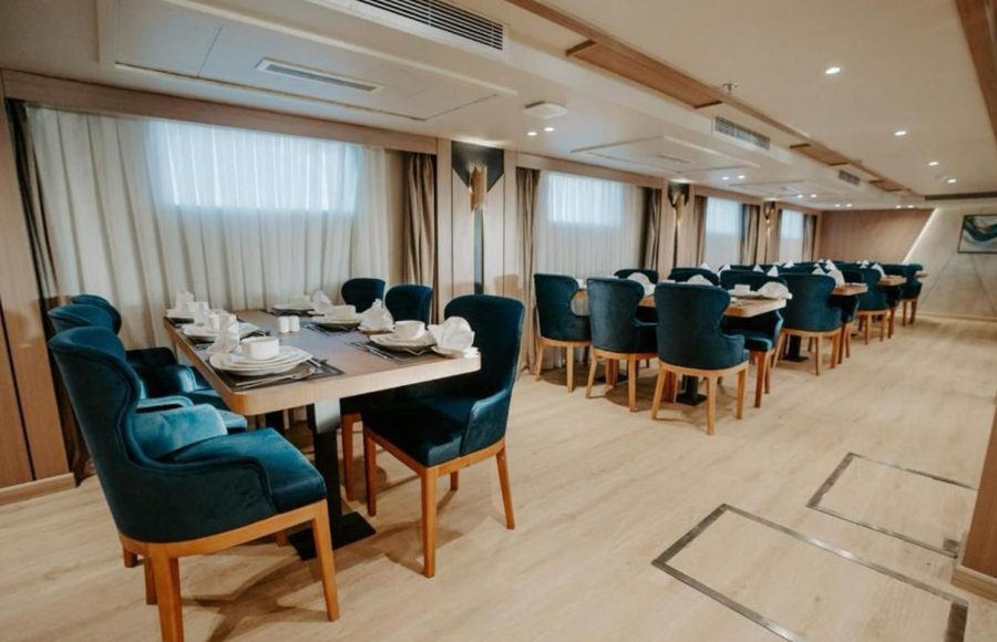 The elegant dining area of a Nile Cruise ship, featuring multiple wooden tables set with white china and paired with deep teal or royal blue velvet upholstered dining chairs.