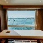 A massage table inside a Nile Cruise spa with a large window view of the Nile River, showing blue water and several white-sailed feluccas (traditional Egyptian sailboats) gliding past.