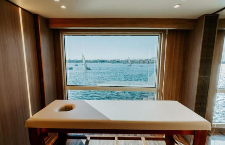 A massage table inside a Nile Cruise spa with a large window view of the Nile River, showing blue water and several white-sailed feluccas (traditional Egyptian sailboats) gliding past.