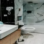 A modern cruise cabin bathroom featuring white marble-look tiles, a glass-enclosed shower/bathtub combo, a white basin sink, and black and white marble accents.