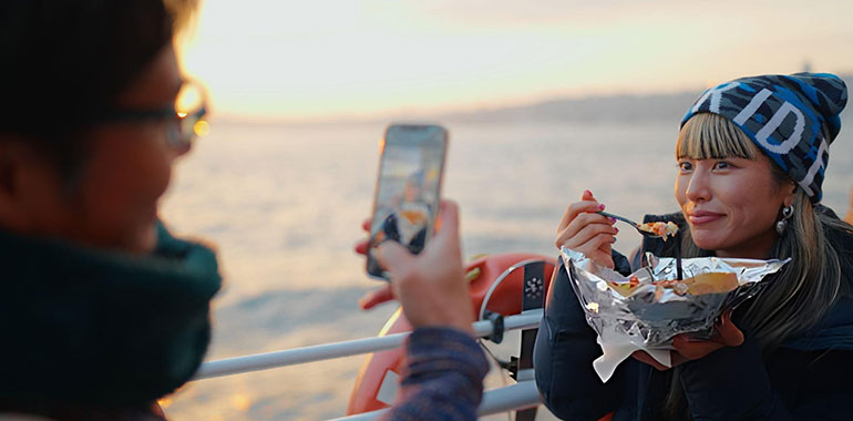 Couple tourist enjoying street food on ferry and capturing memories with smartphone