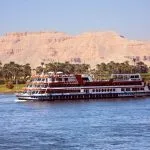 A large, multi-deck river boat is Cruising on the Nile near to Luxor, with green palms lining the riverbank and the rocky, tan-colored sahara desert mountains rising in the background.