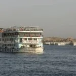 A multi-deck Nile cruise ship, is seen Cruising the Nile, with the ancient Kom Ombo Temple visible on the riverbank in the background.