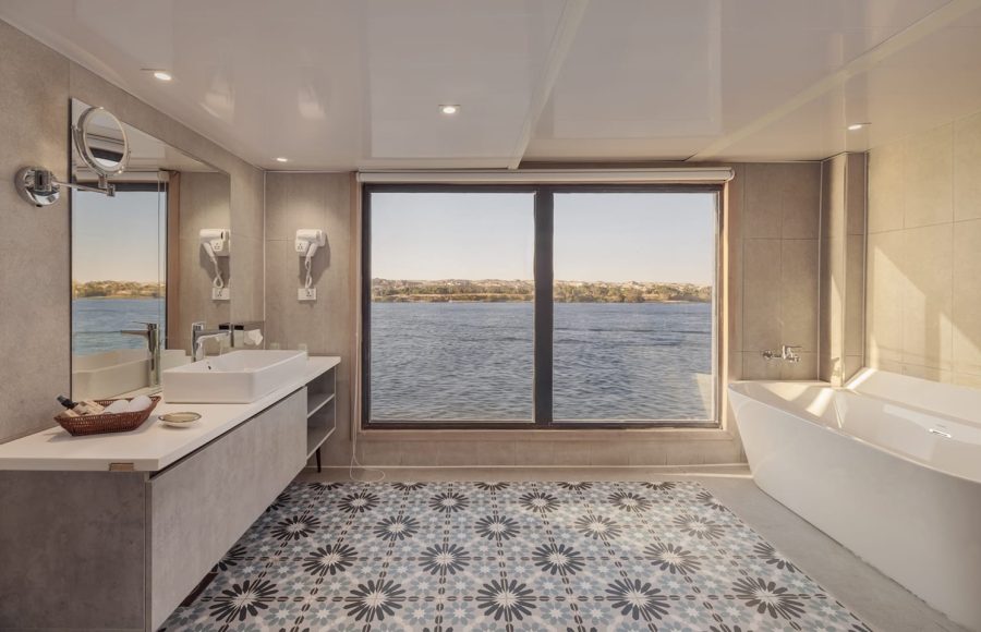 Luxury cruise ship bathroom with a soaking tub, dual sinks, and a large window offering a panoramic view of the Nile River.