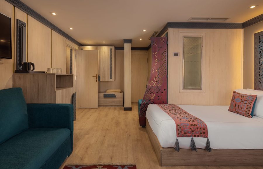Interior of a spacious Nile cruise ship bedroom with a bed, a teal sofa, and patterned bedding, decorated with Egyptian motifs.