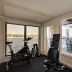Interior of a cruise ship gym with exercise bike and weights, offering a large window view of the Nile River and an adjacent outdoor deck.