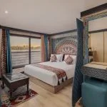 Interior of a luxury cruise ship bedroom suite with a large bed, colorful Egyptian-style wooden headboard, and a panoramic window overlooking the Nile.
