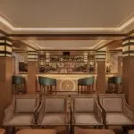 Interior of a sophisticated cruise ship lounge and bar with wood paneling, velvet barstools, and comfortable armchairs.