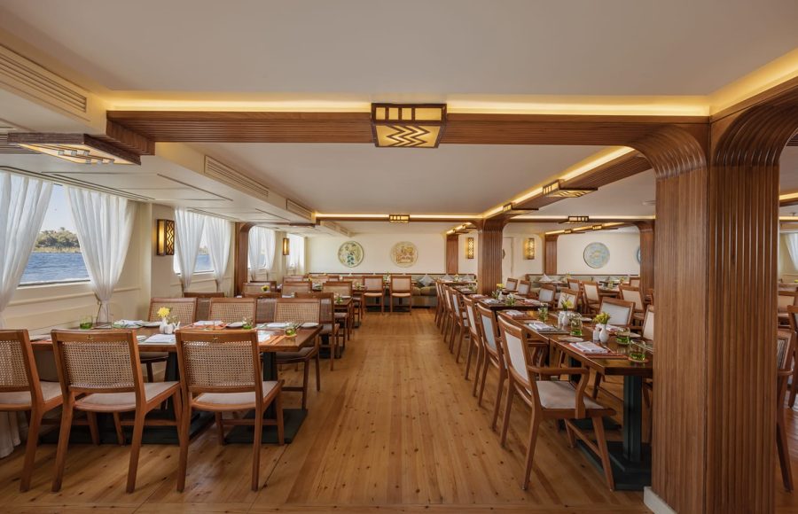 Interior of a long, spacious cruise ship dining room with rows of set tables and carved wooden paneling along the ceiling and walls.
