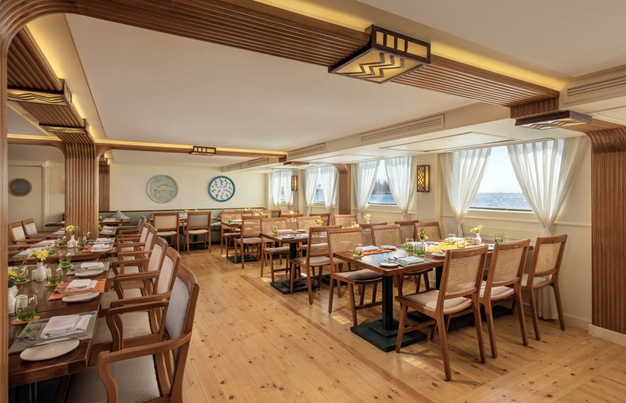 Interior of a bright, contemporary cruise ship dining room with rows of tables, wooden chairs, and large windows overlooking the water.