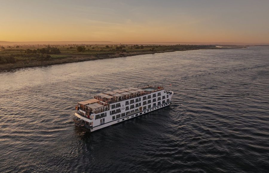 Aerial view of a large, white Nile cruise ship sailing down the river at sunset, with cultivated green fields lining the distant bank.