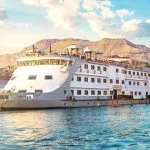 Broadside view of a white and gray Nile cruise ship with a distinctly curved bow, sailing on the river with desert mountains in the background.