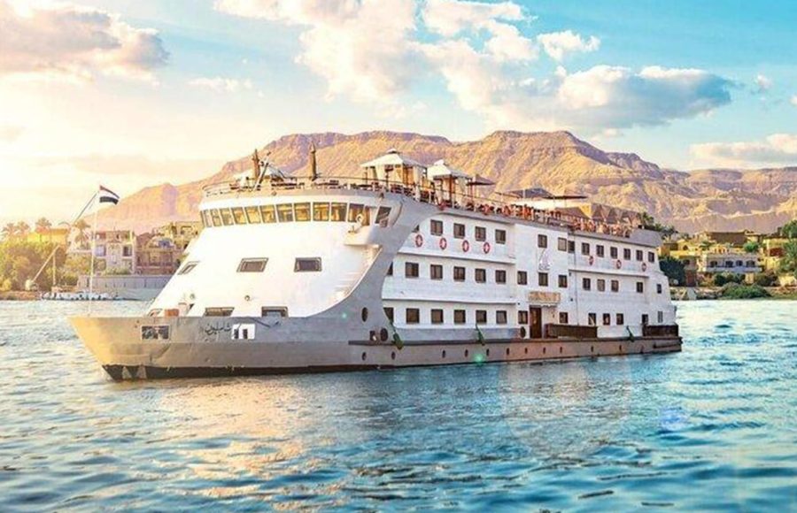 Broadside view of a white and gray Nile cruise ship with a distinctly curved bow, sailing on the river with desert mountains in the background.