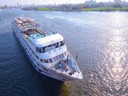 Aerial view of a large, white, multi-deck Nile River cruise ship sailing on the blue water near a green riverbank and city skyline.