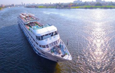 Aerial view of a large, white, multi-deck Nile River cruise ship sailing on the blue water near a green riverbank and city skyline.