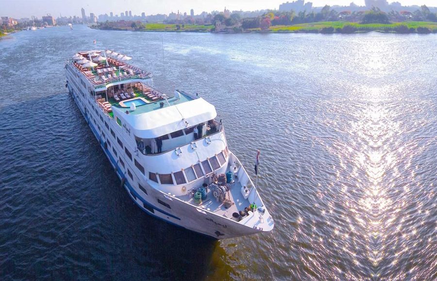 Aerial view of a large, white, multi-deck Nile River cruise ship sailing on the blue water near a green riverbank and city skyline.