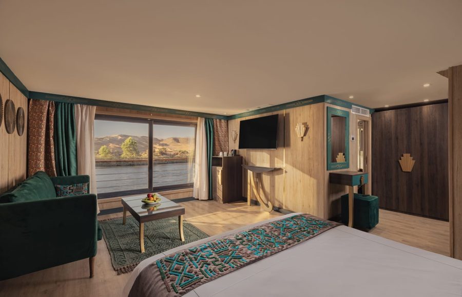 Interior of a luxury Nile cruise ship suite with a large bed, seating area, mounted TV, and a panoramic window view of the river and desert.