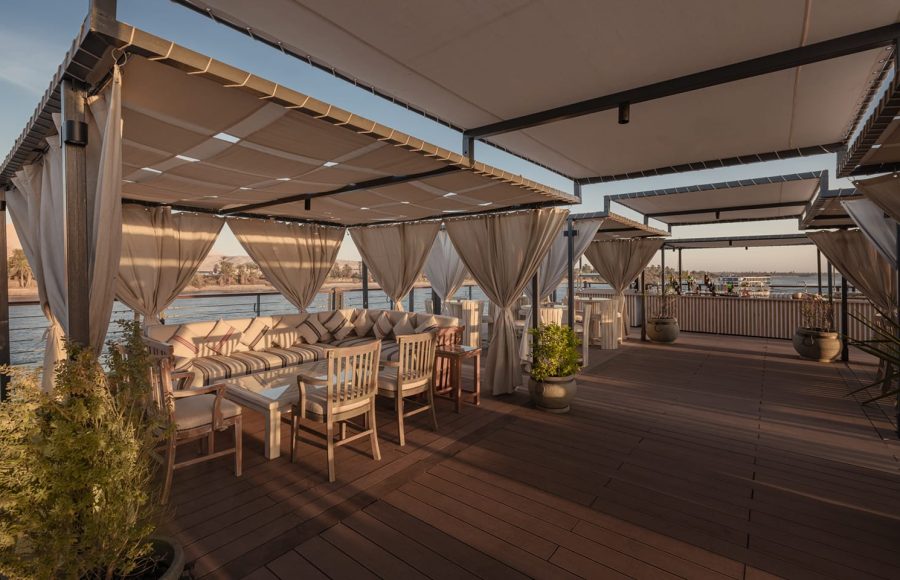 Covered outdoor lounge area on a Nile cruise ship deck with sofas, chairs, and sheer curtains overlooking the river.