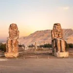 The Egypt Luxor The Colossi of Memnon two massive stone statues of Pharaoh Amenhotep standing against a sunset over the Theban Necropolis hills. The ancient statues serve as a powerful introduction to in depth culture Egypt local tours.