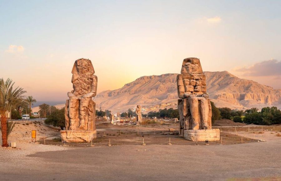 The Egypt Luxor The Colossi of Memnon two massive stone statues of Pharaoh Amenhotep standing against a sunset over the Theban Necropolis hills. The ancient statues serve as a powerful introduction to in depth culture Egypt local tours.