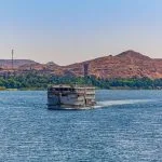 A Cruise boat on the Nile river between Aswan and Luxor Egypt with a view of an Egypt Sunrise at the Nile cruise, showcasing the vessel on the wide river with lush green banks and desert mountains in the background.