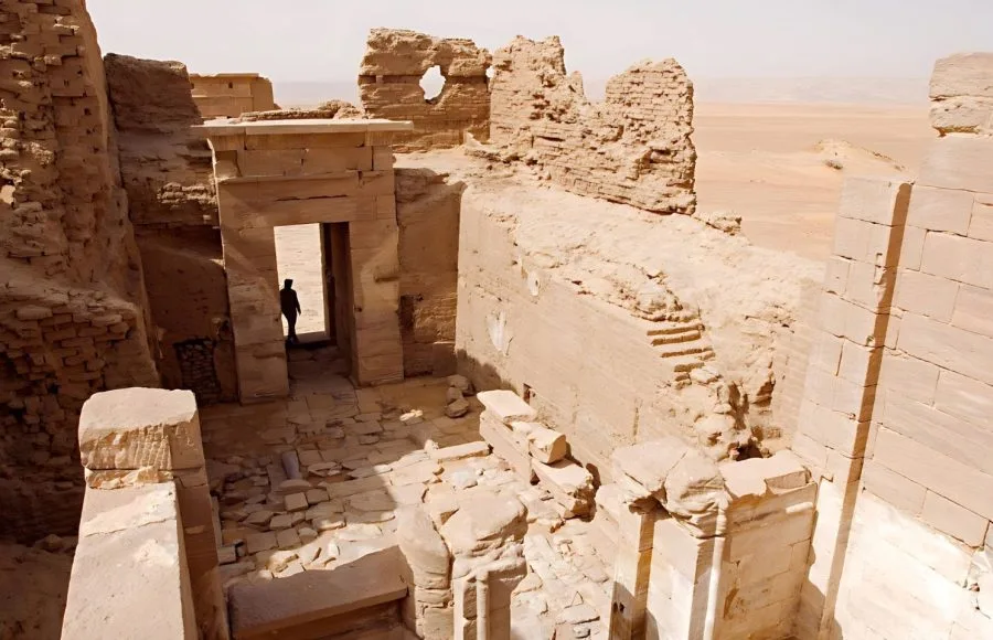 The ruins of the Egypt Temple Of El Dush Baris Oasis, showing crumbling brick walls, doorways, and a single figure standing in the doorway against the desert backdrop.
