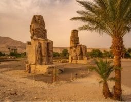 Colossi of Memnon on an Egypt tour from Delhi. The Colossi of Memnon with palm trees and mountains in the background, a sight on an Egypt tour from Delhi.