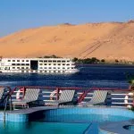 View from a cruise ship's deck showing the pool and lounge chairs, overlooking the Nile River near Aswan with other boats of cruising on the Nile and a barren, sandy hill in the background.
