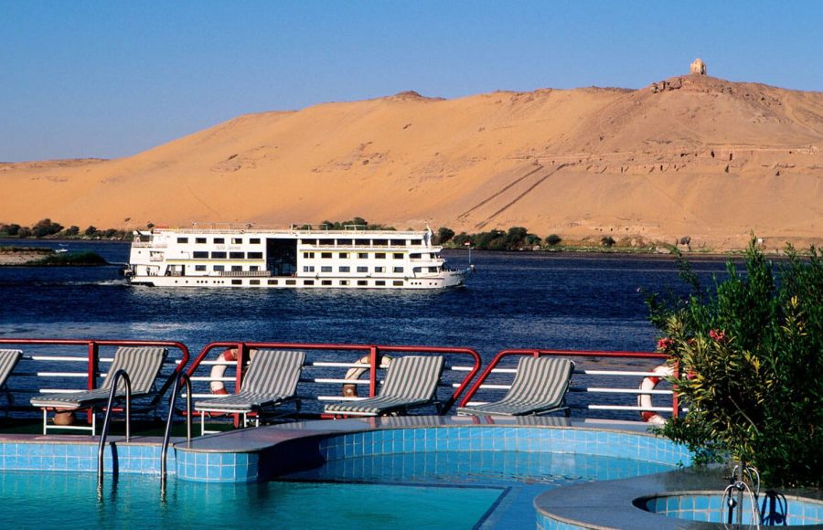 View from a cruise ship's deck showing the pool and lounge chairs, overlooking the Nile River near Aswan with other boats of cruising on the Nile and a barren, sandy hill in the background.