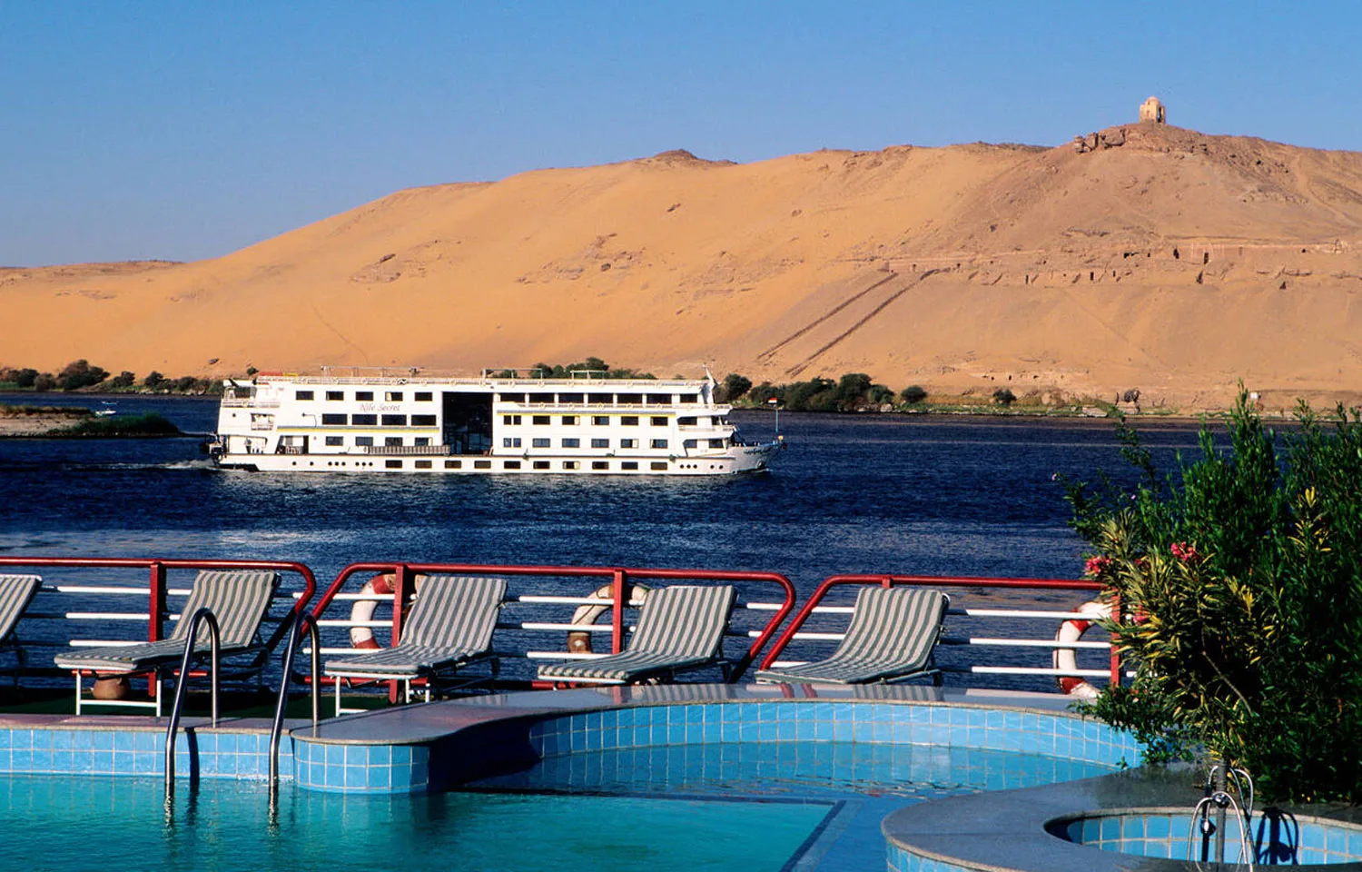 View from a cruise ship's deck showing the pool and lounge chairs, overlooking the Nile River near Aswan with other boats of cruising on the Nile and a barren, sandy hill in the background.