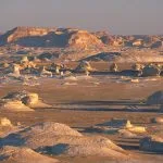 A wide, panoramic view of the Egypt Western Desert White Desert formations, showing numerous white chalk rock outcrops against a tan desert and distant plateau.