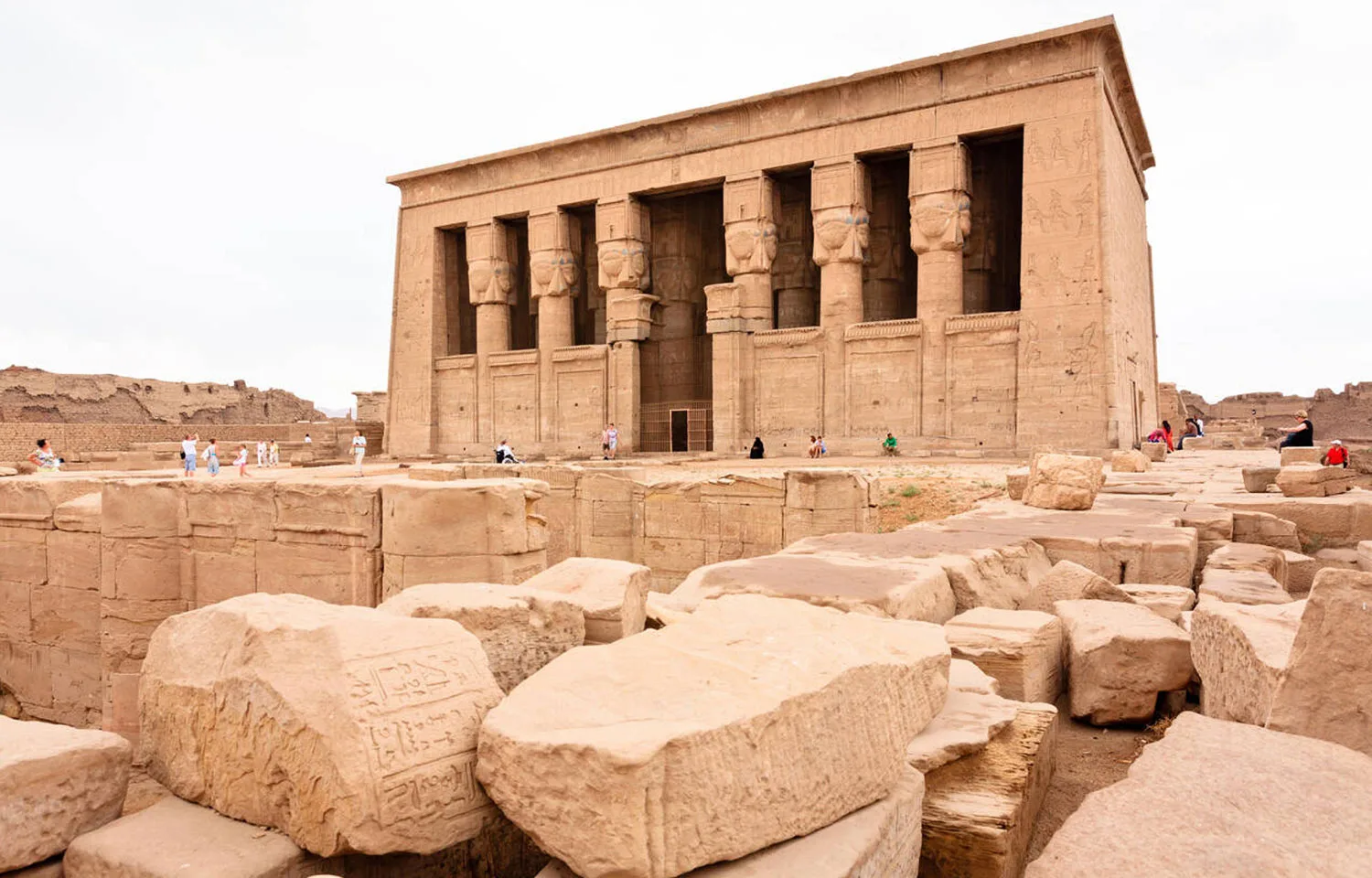 Tourists near the majestic Entrance of Dendera Temple Egypt, showing the massive facade with large columns carved with Hathoric heads, surrounded by fallen stone blocks.