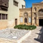 Exterior view of the Coptic Museum in Cairo Egypt, showing the museum's traditional courtyard architecture, a mosaic fountain, and a blend of stone and mashrabiya windows.