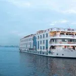 White, multi-deck Nile cruise ship with arched windows sailing on the river at dusk.