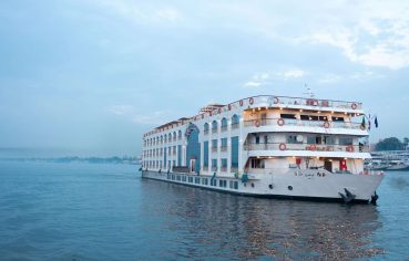 White, multi-deck Nile cruise ship with arched windows sailing on the river at dusk.