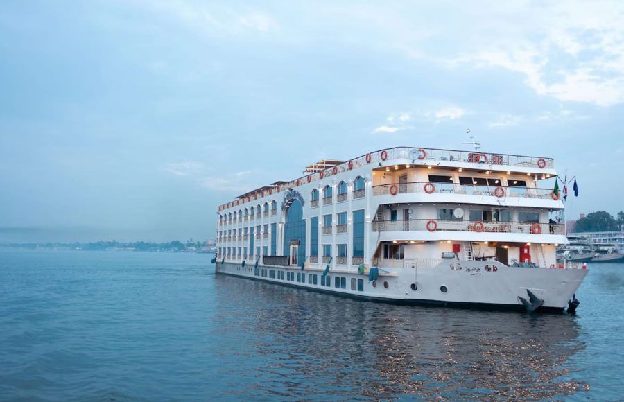White, multi-deck Nile cruise ship with arched windows sailing on the river at dusk.