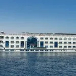 White, multi-deck Nile cruise ship with arched windows sailing in clear blue daylight.