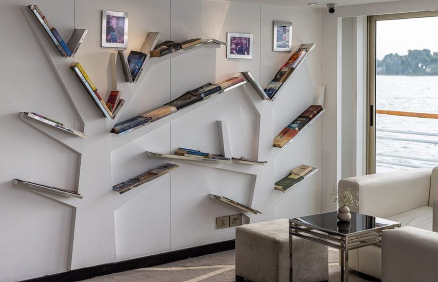 Creative bookshelf installation in the MS Farah library/lounge area, with white sofas and a glass door leading to the deck overlooking the river.