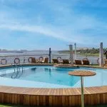 Circular swimming pool on the sun deck of the MS Farah cruise ship, surrounded by a wooden deck and loungers, with a view of the Nile River and landscape.