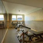 Cruise ship gym with weights, exercise bench, and porthole windows overlooking the water.
