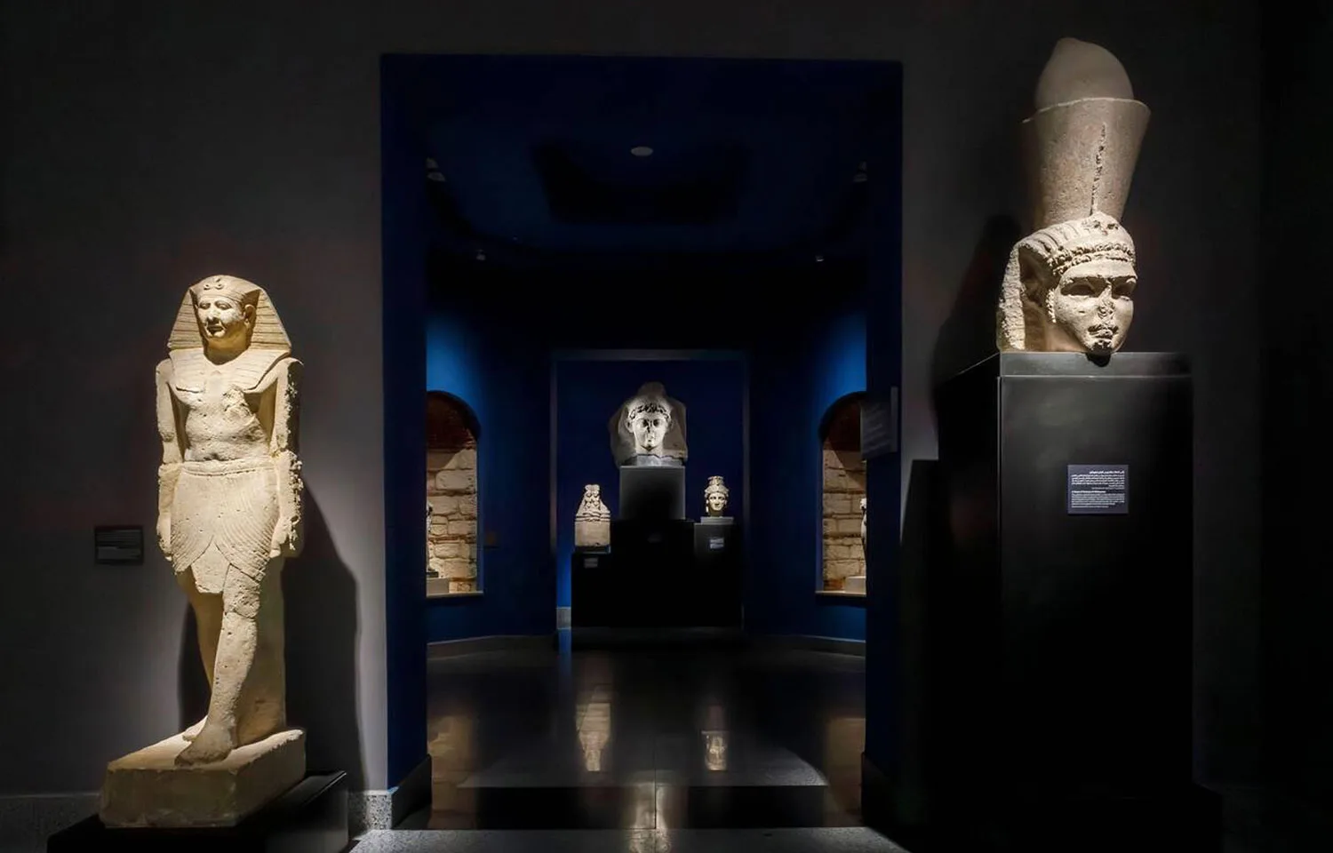 The dimly lit interior gallery of the Greco Roman Museum in Alexandria, Egypt, displaying ancient statues and artifacts under dramatic blue lighting.
