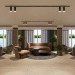 A spacious and elegant lounge on the Hansa Spirit Nile cruise ship, featuring several seating areas with curved sofas upholstered in green and burnt orange velvet. Each area includes dark round coffee tables and accent chairs, centered on dark patterned circular rugs. The space has light wood flooring, black track lighting, and a row of large windows offering views of the Nile and shoreline.